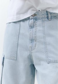 Short en jean bleu clair avec de grandes poches avant et une fermeture par bouton métallique, porté avec une chemise blanche rentrée.