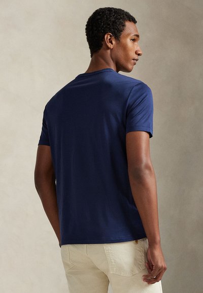 Άνδρας πλευρικά με φανερό μπλε κοντομάνικο πουκάμισο και ανοιχτό μπεζ παντελόνι μπροστά σε ουδέτερο φόντο.