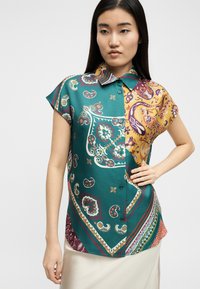 Jeune femme portant un chemisier à manches courtes boutonné avec un motif cachemire en bleu sarcelle, jaune et violet, debout les mains sur les hanches devant un fond blanc.