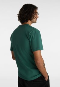 Camiseta verde de manga corta hecha de tela suave, con cuello redondo y un ajuste relajado. Tatuaje visible en el brazo.