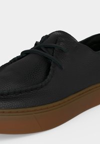 Μαύρο casual παπούτσι από δερματίνη με υφή, ραφές moc toe και σκούρα κορδόνια, επάνω σε χοντρή καφέ λαστιχένια σόλα.