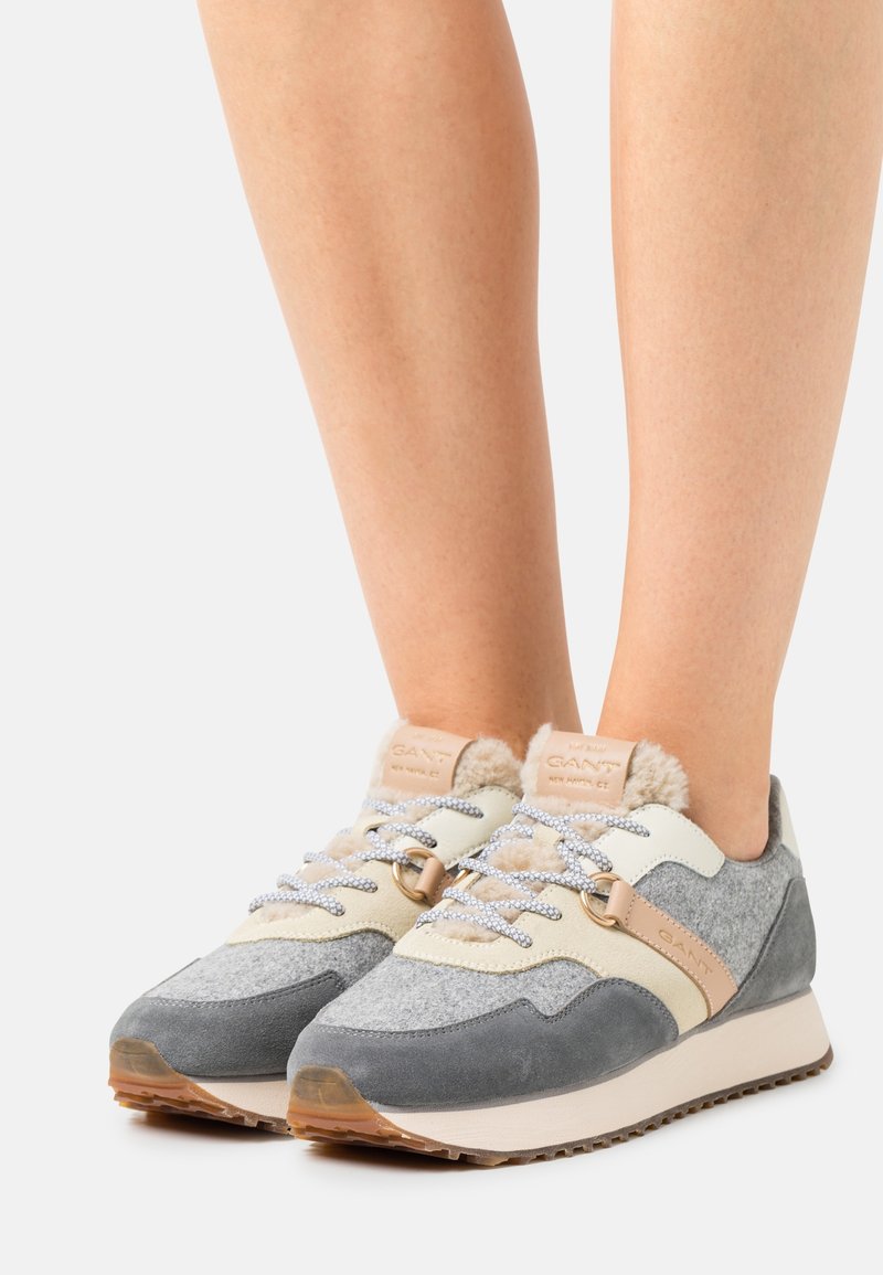 Des pieds portant des baskets GANT grises et beiges avec doublure en fourrure, lacets à motif et semelles en caoutchouc sur un fond blanc.