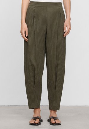 Persona che indossa pantaloni a righe verde oliva e sandali scuri, in piedi su un pavimento chiaro, mani rilassate ai lati, visibile dalla vita in giù.
