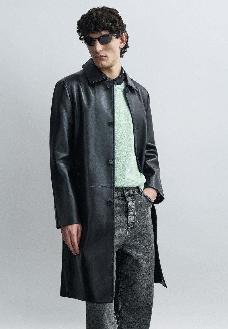 Mees, kes kannab musta nahktagi, mündirohelist kampsunit, halli teksapükse, musta särk ja musti päikeseprille, seisab, üks käsi taskus.