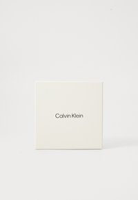 Bela kvadratna škatla z mat površino, na kateri je v črni barvi na sredini spredaj natisnjen "Calvin Klein". Minimalistična zasnova.