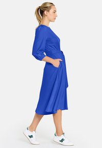 Žena kráčajúca v kráľovskej modrej midi šatách s 3/4 rukávmi a opaskom, doplnená bielymi teniskami s zelenými akcentmi.