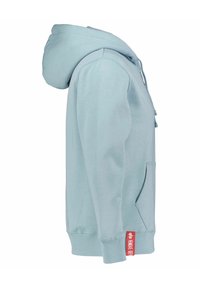 Sweat à capuche bleu clair avec poche avant et étiquette rouge sur le côté inférieur affichant un texte blanc et un petit logo.