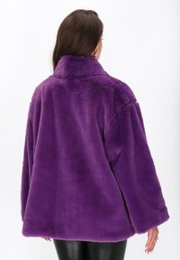 Femeie îmbrăcată cu o jachetă din blană artificială de un violet vibrant, lejeră, văzută din spate, asortată cu pantaloni negri, pe un fundal simplu.