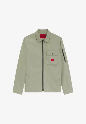 Veste zippée gris clair avec col pointu, poche à rabat sur la poitrine avec bouton-pression, étiquette rouge "HUGO" et poche zippée sur la manche gauche.