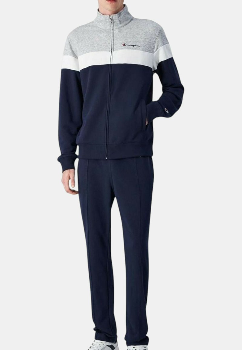 Tuta da allenamento navy con design a blocchi di colore grigio e bianco. La giacca presenta un colletto alto e una zip; i pantaloni hanno una vita elasticizzata e una texture liscia.