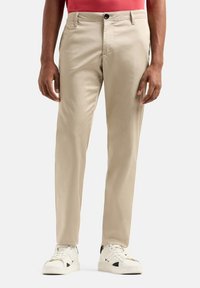 Pantaloni beige con vestibilità sartoriale, dotati di chiusura a bottone e due tasche frontali, abbinati a sneaker bianche e una maglietta rossa.