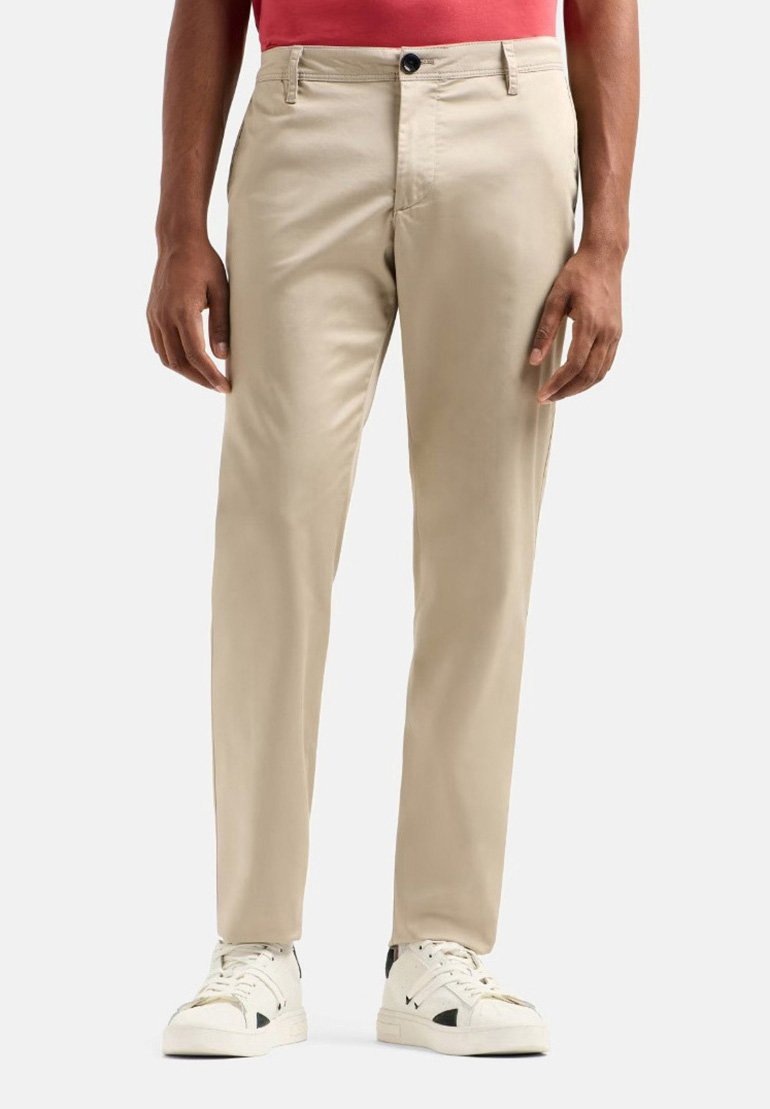 Pantaloni beige con vestibilità sartoriale, dotati di chiusura a bottone e due tasche frontali, abbinati a sneaker bianche e una maglietta rossa.
