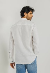 Weißes Leinenhemd mit abgerundetem Saum, langen Ärmeln, knöpfbaren Manschetten und einem Rückenpasse. Kombiniert mit hellblauen Jeans.