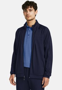 Chaqueta azul marino con cierre de cremallera y cuello alto, superpuesta sobre una camiseta polo azul. Presenta un diseño elegante y detalles sutiles del logotipo.