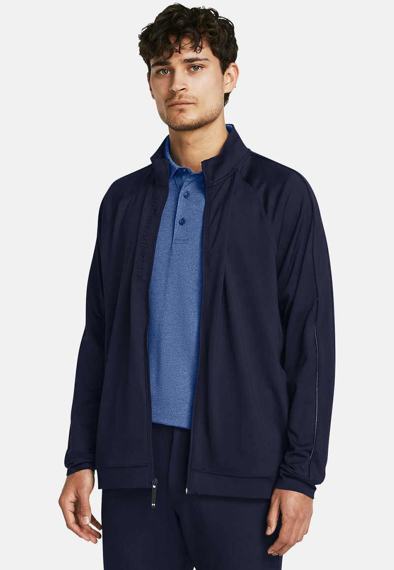 Chaqueta azul marino con cierre de cremallera y cuello alto, superpuesta sobre una camiseta polo azul. Presenta un diseño elegante y detalles sutiles del logotipo.