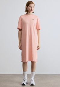 Φόρεμα από βαμβακερό ύφασμα σε ανοιχτό ροζ χρώμα με κοντά μανίκια, πλευρικές σκισμές και μικρό πράσινο λογότυπο στο στήθος, σε συνδυασμό με λευκές κάλτσες και αθλητικά παπούτσια.