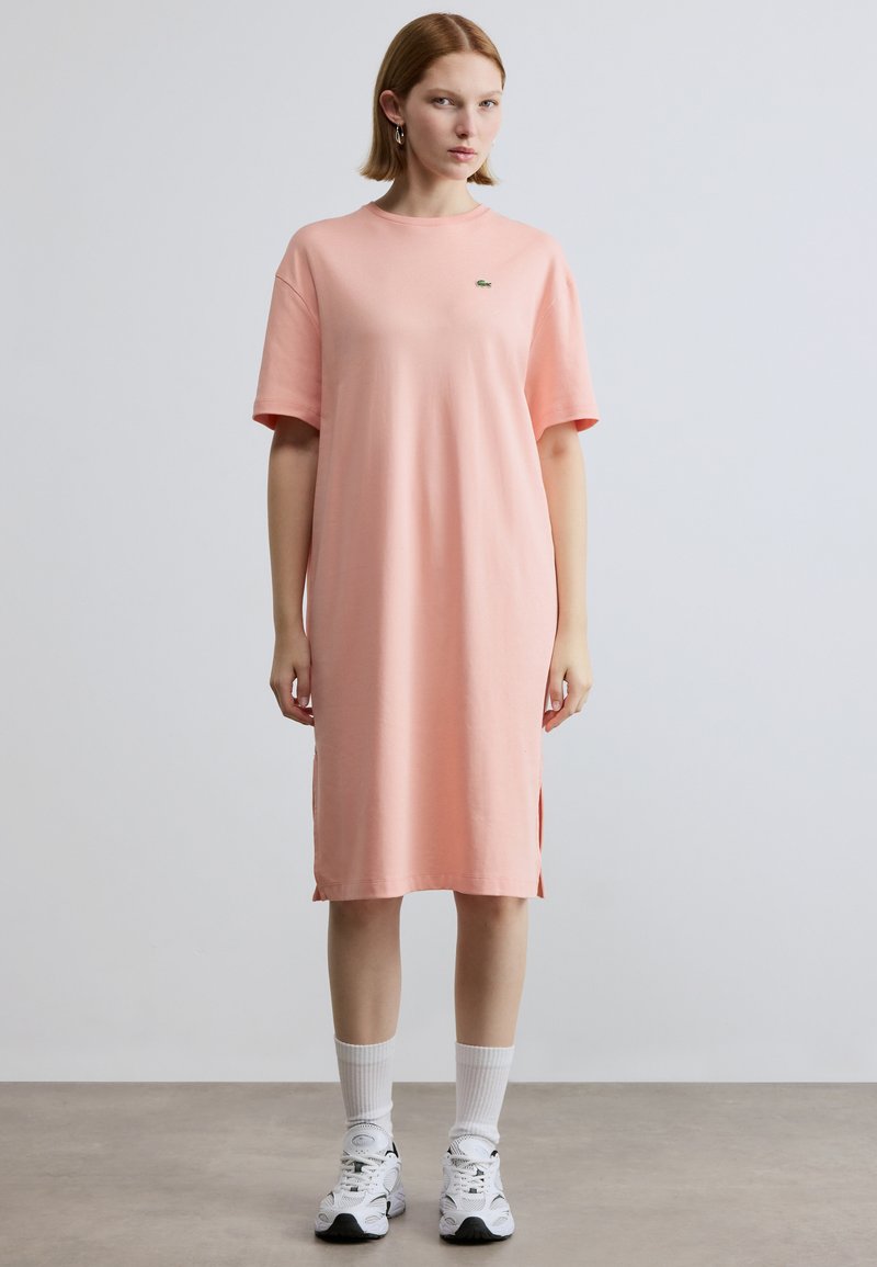 Φόρεμα από βαμβακερό ύφασμα σε ανοιχτό ροζ χρώμα με κοντά μανίκια, πλευρικές σκισμές και μικρό πράσινο λογότυπο στο στήθος, σε συνδυασμό με λευκές κάλτσες και αθλητικά παπούτσια.