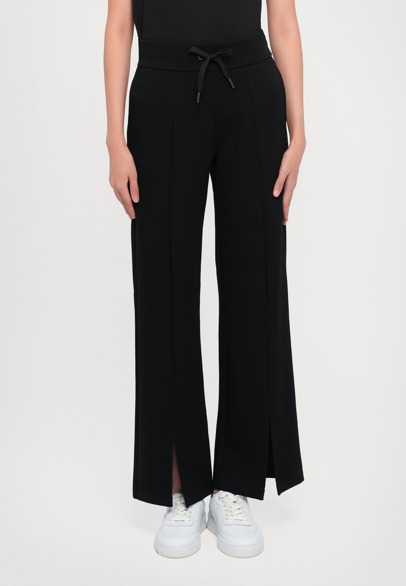 Zwarte wijde broek met een trekkoord in de taille, met zijslits. Gemaakt van een soepele stof, gecombineerd met witte sneakers.