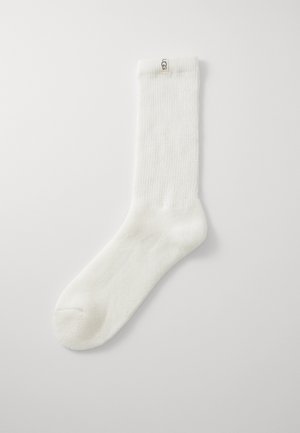 Vit strumpa som når till mitten av vaden, med ribbat mönster och UGG-logotagg högst upp mot en enkel ljus bakgrund.