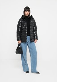 Zwarte gewatteerde jas met een glanzende afwerking, aangesloten silhouet, capuchon en ritssluiting, gecombineerd met lichtblauwe wijde jeans en een zwarte handtas.