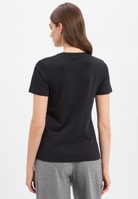 Zwarte katoenen t-shirt met korte mouwen, een ronde halslijn en een gladde textuur, gezien vanaf de achterkant. Grijze broeken zijn gedeeltelijk zichtbaar.
