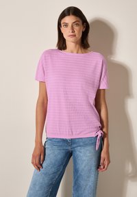 Světle růžový top s krátkým rukávem, který má strukturovaný žebrovaný vzor a detail vázání na boku, spárovaný se světle modrými džíny.