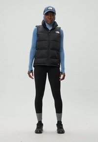 Gilet imbottito nero su una maglietta a maniche lunghe azzurra, leggings neri, calzini grigi e sneakers nere. Indossa anche un cappello abbinato.
