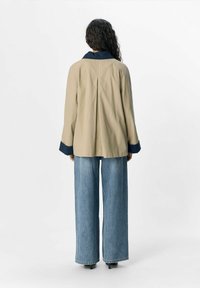 Beige jas met een marineblauwe kraag, voorzien van een plooidetail op de achterkant, gecombineerd met lichtblauwe wijde jeans en zwarte hakken.