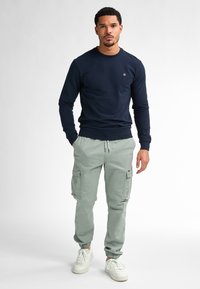 Marineblauwe sweatshirt met lange mouwen en geribbelde manchetten, gecombineerd met lichtgroene cargobroek met zijzakken en elastische manchetten. Witte sneakers maken de look af.