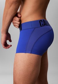 Boxers bleus en tissu extensible. Présente un panneau latéral texturé et une taille avec un logo en noir contrastant.