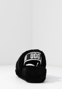 Black fuzzy slipper with a thick sole and a white elastic heel strap branded with "UGG" in bold black letters.