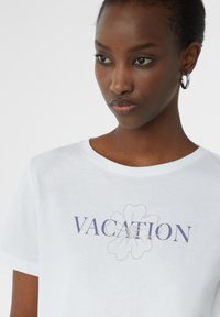 T-shirt en coton blanc avec "VACATION" imprimé en marine, ornée d'un motif floral en perles argentées pour la texture et le détail.