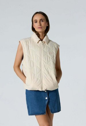 Femme portant un gilet matelassé beige sans manches avec fermeture éclair à l'avant et une jupe en jean bleue avec fente et boutons sur le devant, debout devant un fond uni.