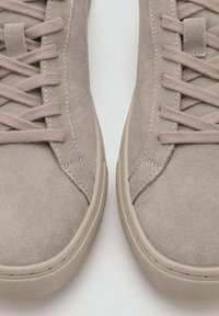 Acercamiento de zapatillas de ante beige con cordones a juego y suelas de goma texturizadas sobre un fondo blanco.