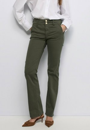 Mujer con pantalones rectos de color verde oliva con cierre de doble botón, camisa blanca abotonada y tacones slingback marrones puntiagudos.