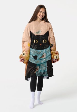 Junge Frau trägt einen übergroßen Hoodie mit großem schwarzen Katzen- und buntem Vogelmotiv, steht vor einem schlichten weißen Hintergrund.