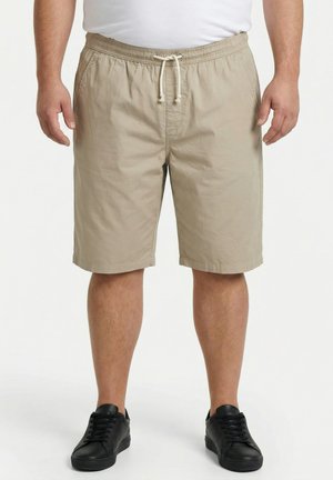 Mand iført beige shorts med snøre, hvid T-shirt og sorte sneakers står foran en ensfarvet hvid baggrund.