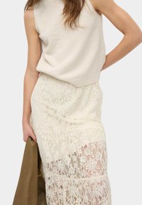 Gebreid mouwloos topje in off-white, gecombineerd met een crèmekleurige kantrok met bloemmotieven en transparante panelen, vastgehouden met een bruine kledingstuk.
