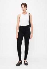 Fekete, skinny fazonú farmernadrág magas derekú, öt zsebes kivitelben, fehér ujjatlan toppal és fekete platform cipővel párosítva, fekete kézitáskával a kezében.