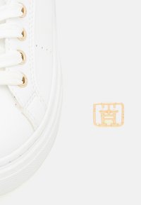 Vita sneakers med strukturerad yta, med guldiga öglor och snören, samt en guldlogotypdetalj på en rektangulär platta.