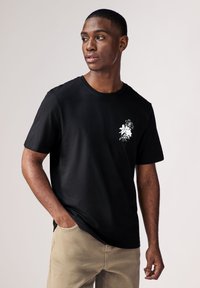 Schwarzes Baumwoll-T-Shirt mit kurzen Ärmeln und Rundhalsausschnitt, mit einem weißen Blumenmotiv auf der linken Brust. Getragen mit beigen Hosen.