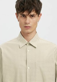 Hele beež värviline nööpidega särk, millel on väike ruuduline muster, klassikaline krae, ühe rinnataskuga ja lihtsate nööpidega. Sile kangapind.