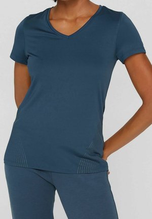 Vrouw draagt een nauwsluitend donkerblauw T-shirt met korte mouwen en V-hals en bijpassende broek, hand op heup tegen een effen achtergrond.