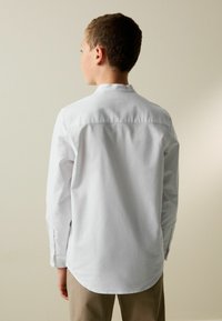Weißes Langarmhemd mit geradem Schnitt, Knopfmanschetten und glatter Stofftextur, von hinten vor neutralem Hintergrund gesehen.
