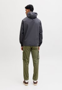 Uomo con giacca con cappuccio grigio scuro, pantaloni cargo verde oliva e sneakers marroni, in piedi di spalle su uno sfondo bianco uniforme.