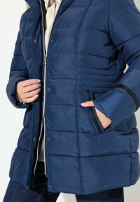 Marinblå puffjacka med quiltat mönster, dragkedja fram och tryckknappar. Har sidofickor med svart kant och ribbade ärmslut.