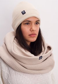 Beige knit beanie with ribbed texture and logo tag, paired with a matching chunky scarf featuring a similar weave and color.