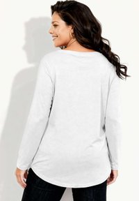 Biała bluzka z długim rękawem, o zaokrąglonym dole i luźnym kroju. Gładka faktura z subtelnym połyskiem, zestawiona z ciemnymi spodniami dżinsowymi.