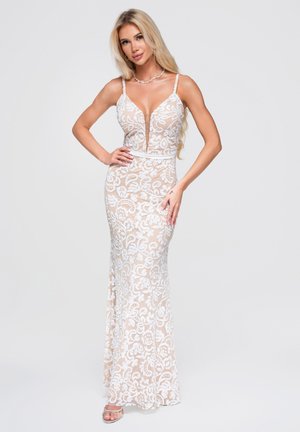 Femme blonde debout dans une robe sans manches en dentelle florale beige et blanche avec un profond décolleté en V et de fines bretelles.