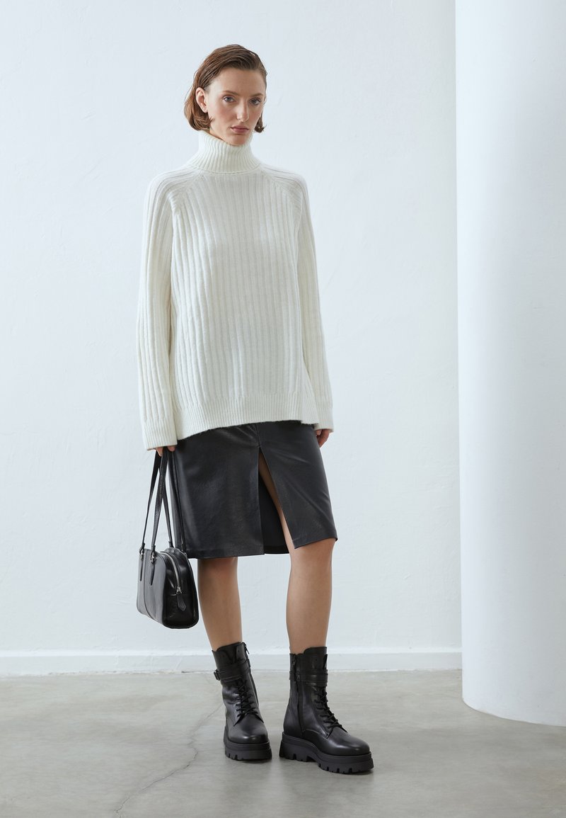 Pull à col roulé blanc surdimensionné à côtes, jupe en cuir noir avec fente à l'avant, bottes noires à lacets et petit sac noir.
